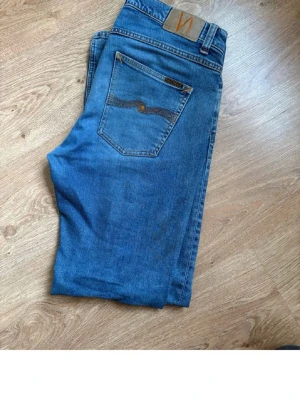 Blå jeans från Nudie Jeans - Märke: Nudie Jeans.                                           Modell: Lost orange Passform: Rak / slim (utifrån bilderna: klassisk rak–slim passform).                Färg: Blå denim Skick: Bra begagnat skick     Snygga Nudie Jeans i klassisk blå tvätt. Tidlös modell som funkar till både vardag och mer uppklätt. Jeansen är i fint skick med naturligt slitage som ger en snygg denimlook. Inga hål eller fläckar. Tillverkade i kvalitetsdenim som håller länge – 
