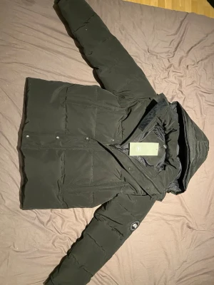 Svart pufferjacka från Canada Goose - Svart pufferjacka från Canada Goose med huva och avtagbar beige pälsdetalj. Jackan har broderad logotyp på ärmen, dragkedja och knappar framtill. Materialet är slitstark polyester och dunfyllning som håller dig varm under kalla dagar. Perfekt för dig som vill ha en stilren vinterlook.