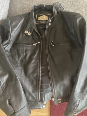 Bikerjacka Livelo - Snygg svart skinnjacka från Livelo med klassisk krage och silverfärgade detaljer. Jackan har två dragkedjeförsedda bröstfickor och knappar vid kragen och nederkant. 