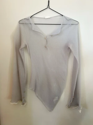 Ljusgrå ribbad topp från Acne Studios - Säljer en superfin ljusgrå ribbad topp från Acne Studios. Toppen har lång ärm, v-ringning med dekorativa silverknappar och asymmetrisk nederkant. Materialet är tunt och stretchigt, vilket ger en snygg passform. Perfekt till jeans eller kjol för en clean look.