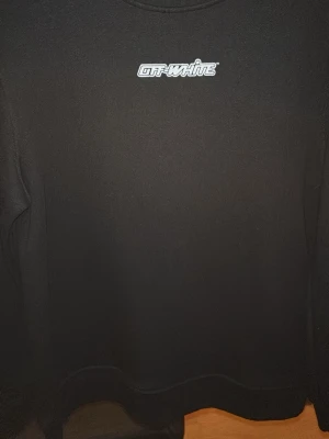 Svart sweatshirt från Off-White - Säljer en svart sweatshirt från Off-White med vit logga på bröstet och stort rött grafiskt tryck på ryggen. Tröjan har rund hals och långa ärmar. Perfekt för dig som gillar streetwear och vill sticka ut med en ikonisk design. AK Pia 