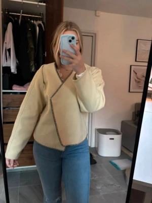 Beige omlottjacka från Vero Moda - Snygg beige omlottjacka från Vero Moda med svarta dekorativa sömmar längs kanterna. Jackan har lång ärm, knappar framtill och en rak, lite boxig passform. Perfekt för dig som gillar stilrena och trendiga plagg.