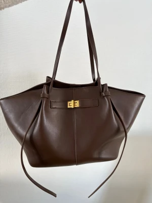 Stor brun shoppingväska i skinn nyskick  - Snygg och rymlig shoppingväska i mörkbrunt skinn med långa axelband och guldfärgat spänne framtill. Väskan har en stilren design med mjuka former och dekorativa remmar på sidorna. Perfekt för dig som vill ha plats för allt du behöver under dagen.