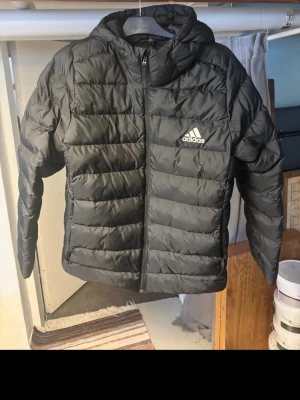 Svart pufferjacka från Adidas - Svart pufferjacka från Adidas med huva och dragkedja framtill. Jackan har ett quiltat mönster och Adidas-logga på bröstet. Tillverkad i polyester och har en normal passform. Perfekt för kyliga dagar när du vill vara både varm och stilren.