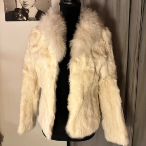 Krämvit pälsjacka med fluffig krage - Säljer en krämvit pälsjacka med extra fluffig krage och långärmad design. Jackan har en mjuk och lyxig känsla, perfekt för dig som vill sticka ut med en cool och trendig look. Passformen är normal och jackan är helfärgad utan mönster.