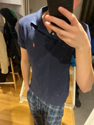Mörkblå pikétröja från Ralph Lauren - Säljer en mörkblå pikétröja från Ralph Lauren med klassisk krage och två knappar framtill. Tröjan har korta ärmar och den ikoniska orangea Polo-loggan broderad på bröstet. Perfekt för en clean och sportig look.