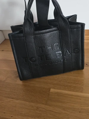 Svart The Tote Bag från Marc Jacobs - Säljer en svart The Tote Bag från Marc Jacobs i präglat skinn. Väskan har både handtag och en bred axelrem, samt präglad logga framtill. Insidan har ett stort fack och en innerficka med dragkedja. Perfekt för dig som vill ha en stilren och rymlig väska. Fint skick