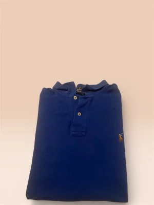 Blå pikétröja från Polo Ralph Lauren - Snygg blå pikétröja från Polo Ralph Lauren med klassisk krage och två knappar framtill. Tröjan har det ikoniska broderade polospelaren på bröstet i brunt, vitt och blått. Tillverkad i mjuk bomull som är skön mot huden.