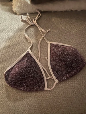 Glittrig lila bikiniöverdel - Säljer en snygg trekantsformad bikiniöverdel i glittrigt lila tyg med vita kanter och knytband i nacken och ryggen. Perfekt för dig som vill sticka ut på stranden eller vid poolen!