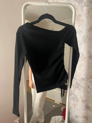 Svart ribbad långärmad topp - Säljer en svart långärmad topp med ribbad struktur och snörning i sidan. Toppen har en asymmetrisk halsringning och är croppad i modellen. Perfekt för dig som gillar stilrena plagg med en twist.