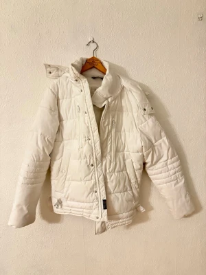 Vit pufferjacka med huva - Säljer en vit pufferjacka med avtagbar huva och quiltad design. Jackan har dragkedja och knappar framtill, flera fickor med dragkedja och snygga detaljer vid midjan. Perfekt för kalla dagar och har en modern look som passar till streetstyle.