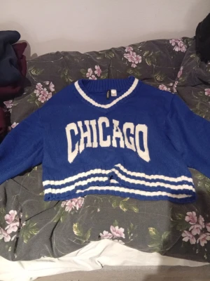 Blå croppad stickad tröja Chicago H&M - Snygg blå croppad stickad tröja från H&M Divided med vit text 'CHICAGO' framtill. Tröjan har vita ränder vid halsringning och nederkant, samt långärmad och ribbad design. Perfekt för dig som gillar sportig och avslappnad stil.