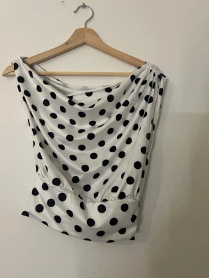 Vit prickig omlott topp - Säljer en vit omlott topp med svarta prickar. Toppen har en lös passform upptill och sitter tightare i midjan, vilket ger en snygg siluett. Den är ärmlös och kan bäras offshoulder för en cool look. Materialet känns mjukt och stretchigt, perfekt för att styla på olika sätt.