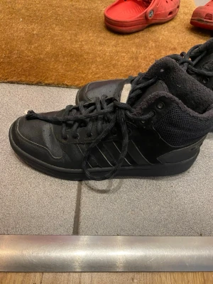 Svarta höga sneakers från Adidas - Säljer ett par svarta höga sneakers från Adidas med klassiska tre ränder på sidan. Skorna har snörning, rund tå och är tillverkade i en mix av skinn och textil med meshdetaljer. Insidan är fodrad för extra komfort och de har en platt sula.