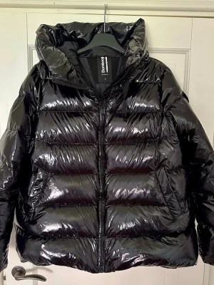 Svart pufferjacka från Bomboogie - Säljer en glansig svart pufferjacka från Bomboogie i storlek 4. Jackan har huva med snörning, dragkedja framtill och två sidofickor. Fyllningen är äkta dun och jackan har en quiltad look som ger extra volym och värme. Perfekt för kalla dagar då tyget även är vattenavvisande.