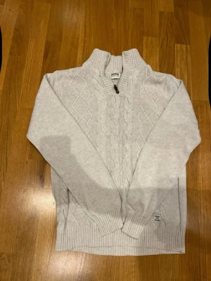 Ljusgrå stickad tröja från Dobber - Säljer en ljusgrå stickad tröja från Dobber i storlek medium. Tröjan har ett snyggt kabelstickat mönster framtill, ribbad krage och muddar samt en liten knappdetalj vid halsen. Perfekt för kyliga dagar och enkel att matcha med jeans.