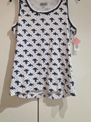 Vit blommig linnetopp från Miss Milla - Supersöt ribbad linnetopp från Miss Milla i vitt med blått blommönster och små svarta prickar. Toppen är ärmlös och har rundad halsringning med kontrastkant. Tillverkad i 100% bomull och perfekt för varma dagar.