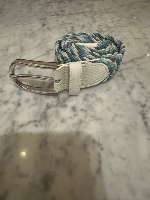Flätad blå och vitt bälte - Säljer ett snyggt flätat bälte i blå, vit  och grå toner med vita detaljer vid spännet och ändarna. Bältet har ett silverfärgat metallspänne och är gjort i ett slitstarkt syntetmaterial. Perfekt för att ge outfiten lite extra stil.