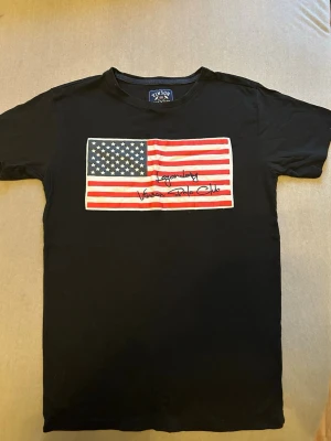 Svart t-shirt Vinson Polo Club S - Svart t-shirt från Vinson Polo Club med stor amerikansk flagga tryckt på bröstet och broderad text. Rund hals och korta ärmar. Materialet är mjuk bomull som känns skön mot huden. Perfekt för dig som gillar streetstyle och vill sticka ut med en cool flaggdesign.