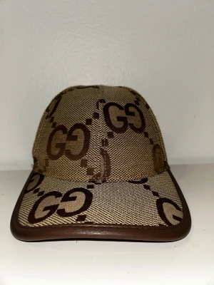 Brun och beige keps - Snygg keps i beige och brunt med en snygg-logga över hela. Tillverkad i bomull och polyester med detaljer i brunt skinn. Justerbar rem baktill med stämpel. Perfekt för dig som gillar lyxiga accessoarer och vill sticka ut.