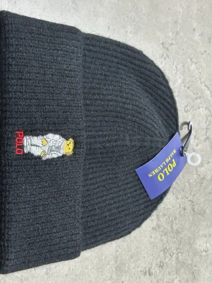 Svart mössa Polo Ralph Lauren - Ribbstickad svart mössa från Polo Ralph Lauren med broderad ikonisk björn på framsidan och Polo-text i rött. Klassisk design med uppvikt kant och mjukt material som håller dig varm.