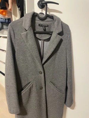 Grå kappa från trf_outerwear, S - Säljer en stilren grå kappa från trf_outerwear i storlek S. Jackan har klassisk krage, två framfickor och stängs med knappar framtill. Materialet är mjukt och har en ull-liknande känsla, perfekt för kyliga dagar.