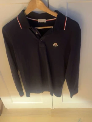Mörkblå långärmad Moncler piké - Snygg mörkblå långärmad piké från Moncler med klassisk krage och knappar framtill. Röd och vit randig detalj på kragen och ärmslut samt ikonisk Moncler-logga på bröstet. Tillverkad i 100% bomull för skön känsla.