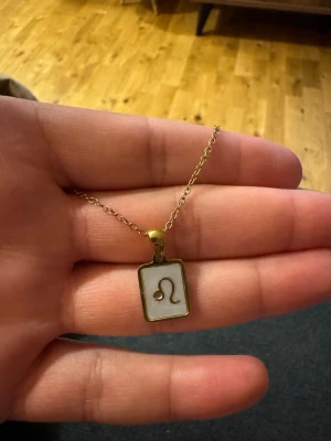Stjärntecken halsband ♌ Lejonet – nyskick - Bilderna visar ett halsband med ett rektangulärt hänge i guld och vitt, dekorerat med Leosymbolen. Ingen väst syns på bilderna.