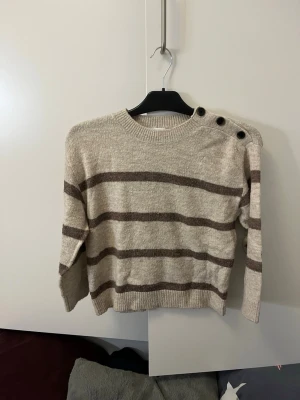 Stickad randig tröja från Vero Moda. Strl: 134/140 - Säljer en stickad randig tröja från vero Moda, med knappar på ena axeln. Beige och brun.
