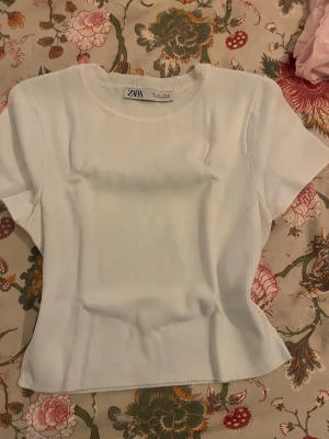Vit ribbad t-shirt från Zara - Säljer en vit ribbad t-shirt från Zara i storlek L. T-shirten har rund hals och korta ärmar, med en slim passform som sitter snyggt på kroppen. Materialet är mjukt och stretchigt, perfekt för en clean och fräsch look.