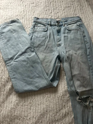 Ljusblå ripped jeans med straight fit - Säljer ett par ljusblå jeans med straight fit och snygga slitningar vid knäna. Klassisk femficksmodell med hög midja och dragkedja. Jeansen har en chill vibe och är gjorda i mjukt denim-material. Perfekt för dig som gillar en avslappnad stil med lite edge.
