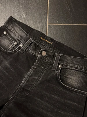 Svarta Nudie Jeans, storlek W30 L32 - Snygga svarta jeans från Nudie Jeans med diskret tvättad look och klassisk femficksdesign. Jeansen har skinny passform, svarta sömmar och Nudie Jeans logga på bakfickan. Perfekt för dig som gillar stilrena och smala jeans.