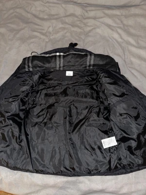 Svart pufferjacka från Burberry - Svart pufferjacka från Burberry med coolt Burberry London England-märke på ärmen. Jackan har dragkedja och tryckknappar framtill, två zip-fickor och en krage med rutigt mönster i grått och vitt. Materialet är polyester och jackan är quiltad för extra värme.