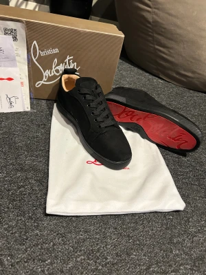 Svarta sneakers Christian Louboutin - Säljer ett par svarta sneakers från Christian Louboutin i mocka med klassisk röd sula och diskret logga undertill. Skorna har rund tå, svarta snören och en stilren, minimalistisk design. Insidan är beige och de kommer med originalkartong och dustbag.