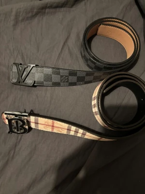 Louis Vuitton & Burberry bälten - Två snygga designerbälten: ett Louis Vuitton i svart och grått med rutigt mönster och stor LV-spänne, samt ett Burberry i beige, brun och röd med klassiskt rutigt mönster och svart B-spänne. Båda är i skinn och perfekta för att lyfta din outfit. Båda är 95 cm långa så passar de flesta 