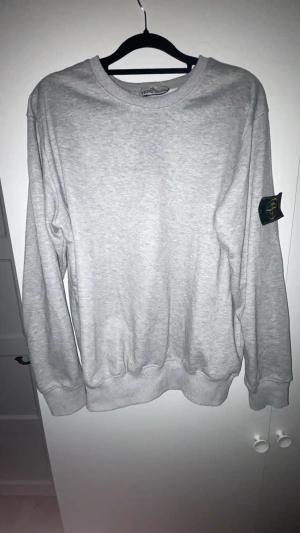 Grå sweatshirt från Stone Island - Snygg grå sweatshirt från Stone Island med klassisk patch på vänster ärm. Tröjan har rund hals, ribbade muddar och är tillverkad i mjuk bomull. Perfekt för dig som gillar stilrena och enkla plagg med streetkänsla. Finns påse till och pris kan diskuteras 