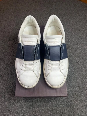 Valentino Garavani vita sneakers - Snygga vita sneakers från Valentino Garavani med bred mörkblå läderdetalj över snörningen och coola nitar på hälen. Skorna har rund tå, platt sula och är tillverkade i skinn. Perfekt för dig som gillar exklusiv streetstyle och italiensk design. Allt OG medföljer inklusive snörre. Prisr diskuterbart vid snabb affär.