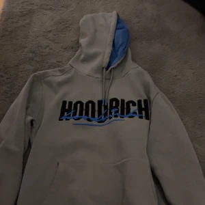 HOODRICH tröja - Säljer en grå hoodie från HOODRICH med stor svart och blå logga på bröstet. Huvan har blått foder och dragsnören. Klassisk känguruficka framtill och mjukt material på insidan. Perfekt för dig som gillar streetwear och vill sticka ut.