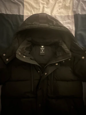 Svart pufferjacka från Smog - Svart pufferjacka från Smog med huva, dragkedja och tryckknappar framtill. Jackan har två bröstfickor med dragkedja och flera tryckknappar längs fronten. Perfekt för kalla dagar med sin tjocka och vadderade design.