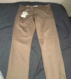 Bruna kostymbyxor från TeeShoppen - Snygga bruna kostymbyxor från TeeShoppen med klassisk rak passform. Byxorna har bälteshällor, knapp och dragkedja fram samt diskreta sidofickor. Materialet är mjukt och har en lätt glansig finish som ger en stilren look.