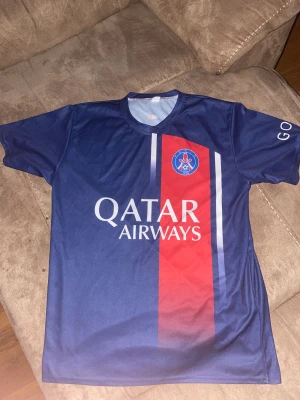 PSG Mbappé #7 blå fotbollströja - Säljer en snygg PSG matchtröja med Mbappé och nummer 7 på ryggen. Tröjan är blå med röd vertikal rand och har Qatar Airways tryck på framsidan. Korta ärmar och tillverkad i lätt polyester, perfekt för fotboll eller häng med kompisar.