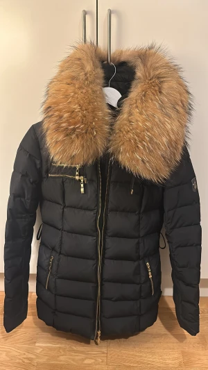 Svart pufferjacka Hollies med päls - Svart pufferjacka från Hollies med stor fluffig brun päls runt kragen och leopardmönstrat foder. Jackan har guldfärgade dragkedjor, flera fickor och snygga detaljer på ärmen. Perfekt för dig som vill ha en varm och trendig vinterjacka med extra attityd.