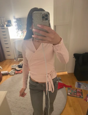 Ljusrosa omlottopp med knyt - Säljer en ljusrosa omlottopp med långa ärmar och snygg v-ringning. Toppen har ett knyt i midjan som ger en feminin siluett och är tillverkad i mjukt bomullsmaterial. Perfekt för dig som gillar stilrena och trendiga plagg med en söt touch. 