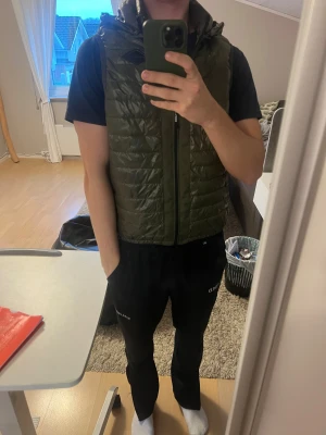 Olivgrön dunväst från Replay - Snygg olivgrön dunväst från Replay med huva och quiltad design. Västen har dragkedja framtill med Replay-logga och ett Replay-märke på bröstet. Perfekt att slänga över en hoodie för extra värme under hösten eller vintern. Storlek M.