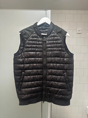 Svart Barbour väst - Svart pufferväst från Barbour International i storlek M. Västen har quiltad design, två fickor med knappstängning och tydlig Barbour-logga på bröstet. Materialet är syntet med fibredown-fyllning och dragkedja framtill. Perfekt för lager-på-lager under höst och vinter.