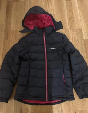 Svart pufferjacka från Everest - Svart pufferjacka från Everest med huva och rosa foder. Jackan har dragkedja framtill, två fickor med dragkedja och diskret logga på bröstet. Perfekt för kalla dagar med sin vadderade design och snygga kontrastfoder. Strl 140