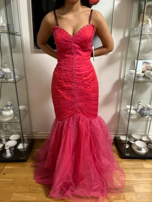 Skrikrosa  långklänning med glitter och volang - Säljer en skrikrosa långklänning utan axelband, figurnära passform och volang nertill. Klänningen har glittriga detaljer och en snygg v-ringning. Materialet är lätt och skimrande, perfekt för bal eller fest. Supersnygg och unik look!