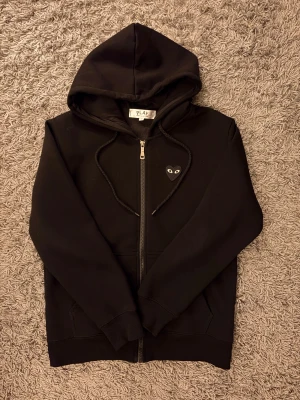 Svart hoodie Comme des Garçons Play - Svart hoodie från Comme des Garçons Play med dragkedja, huva och snörning. Ikoniskt hjärta med ögon på bröstet och stort tryck på ryggen. Tillverkad i mjuk bomull, perfekt för en chill och stilren look.