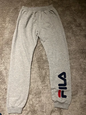 Grå mjukisbyxor från FILA - Snygga grå mjukisbyxor från FILA med stort tryckt logotyp i blått och rött längs benet. Byxorna har resår i midjan och vid bensluten, samt fickor på sidorna. Perfekta för chill eller träning, riktigt sköna och stilrena.