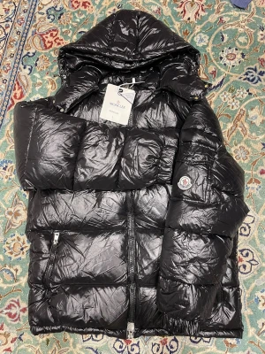 Svart Moncler pufferjacka  - Svart pufferjacka från Moncler med glansig finish, stor huva och klassisk logga på armen. Jackan har dragkedja framtill, fickor med dragkedja och quiltad design. Perfekt för kalla dagar när du vill ha både stil och värme.